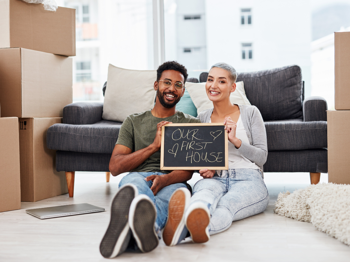 resolutions for home buyers in 2025 photo of 2 first time home buyers sitting in front of a couch holding a sign that reads our first home